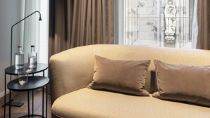 A modern room with a beige sofa, a small side table with bottles, and a view of a gothic-style building through a window with curtains. - Legend Hotel (Cologne)