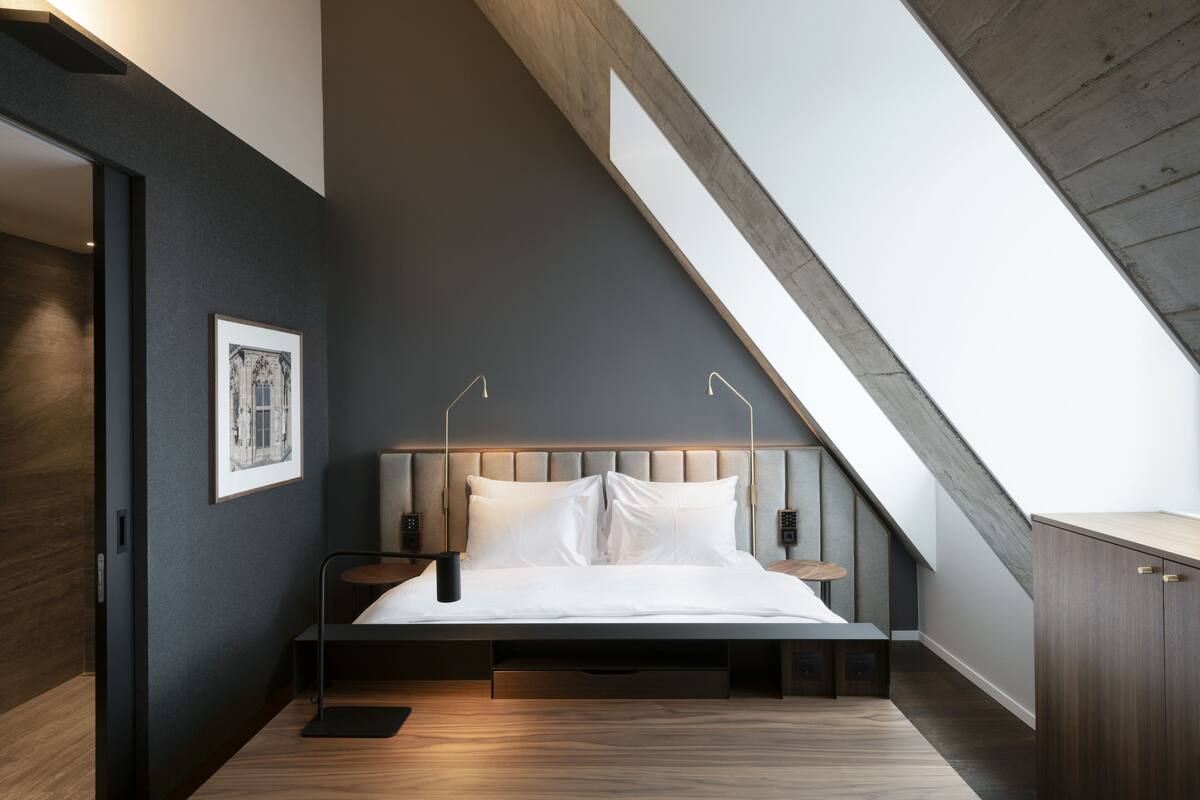 A modern bedroom with a sloped ceiling, a bed with white linens, a framed picture on the wall, and a wooden floor.