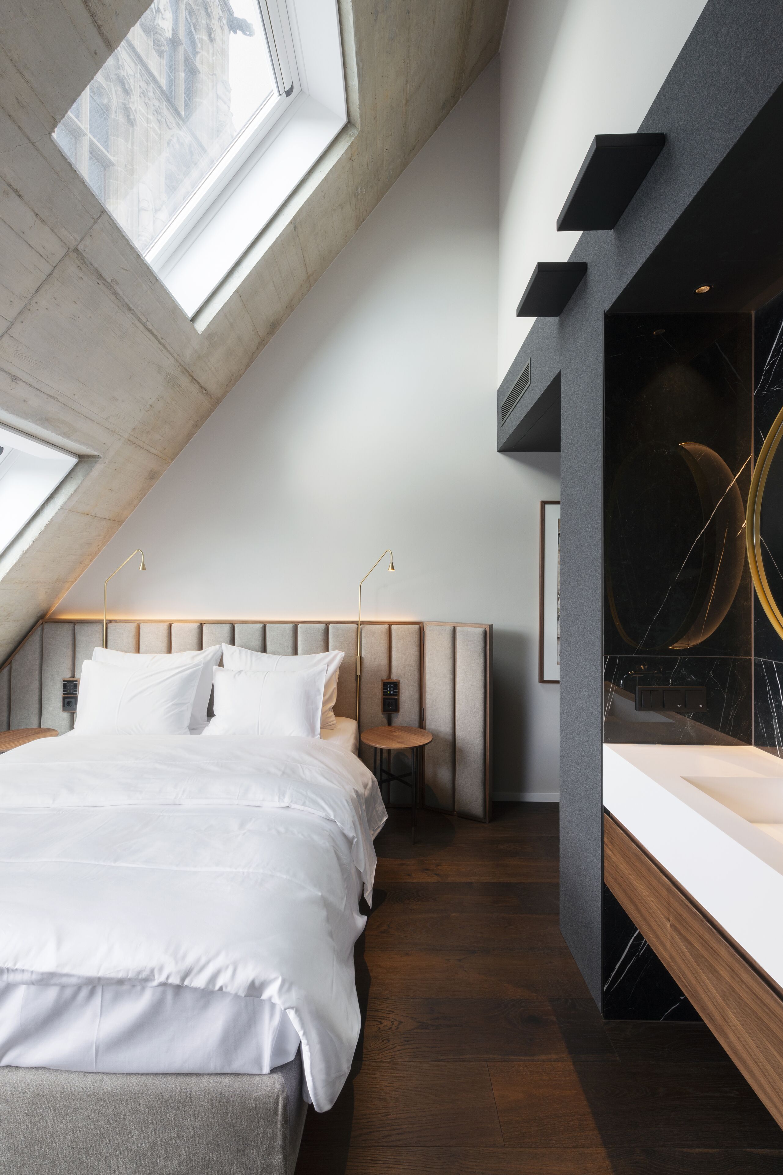 A modern bedroom with a sloped ceiling, skylight, a bed with white bedding, a bedside table, and a vanity with a mirror.