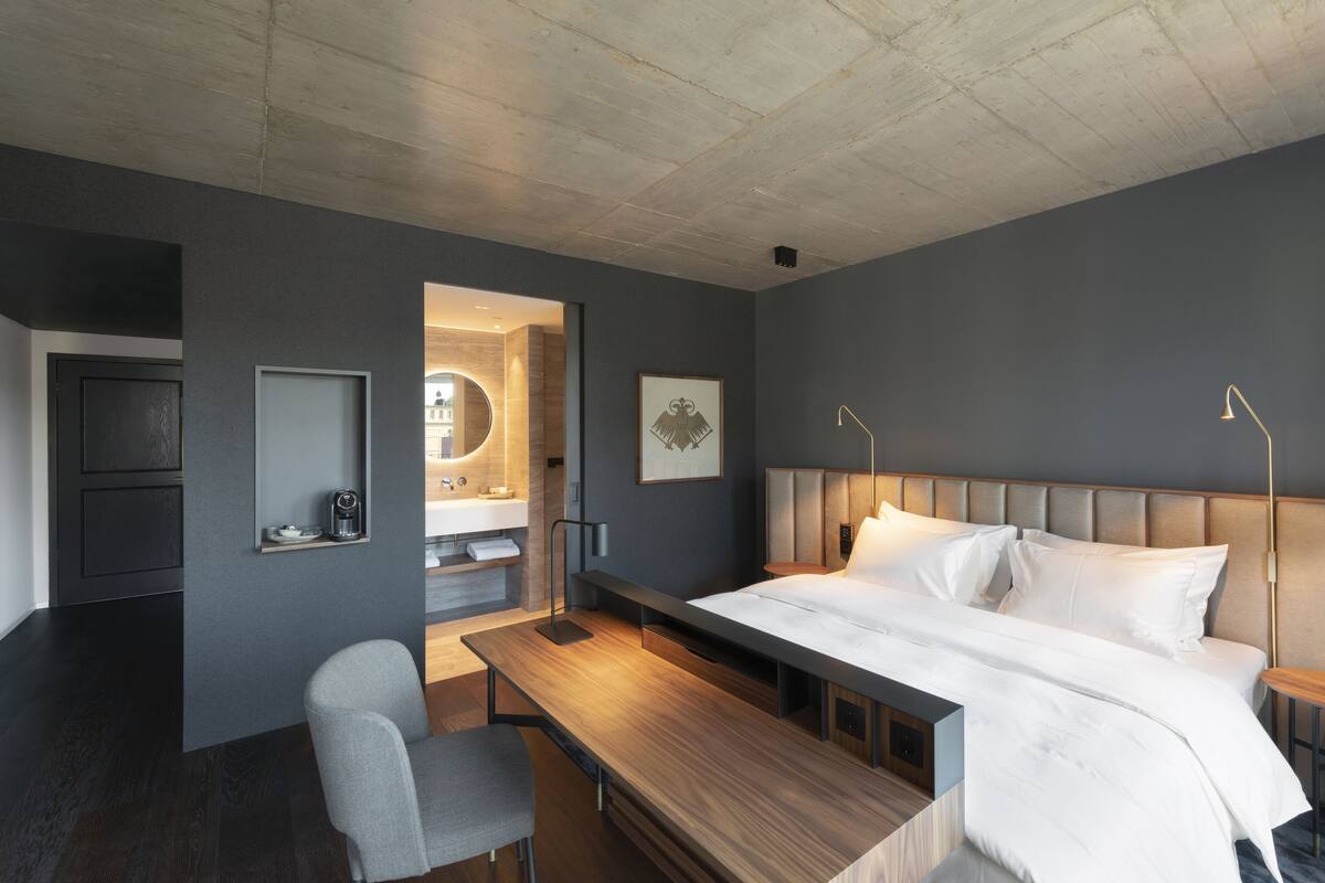 A modern hotel room with a large bed, a desk, a chair, a bathroom with a mirror, and a small seating area.