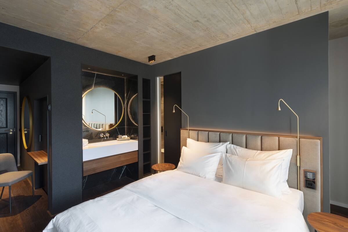 A modern hotel room with a large bed, a wooden desk, a round mirror, and a built-in vanity.