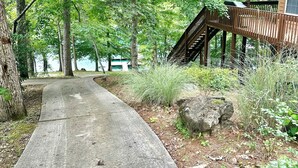 Un chemin de béton mène à une terrasse en bois, accessible par un escalier, et est entouré d’un feuillage verdoyant et dense, avec un grand rocher au premier plan.