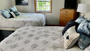 Une chambre avec deux lits, un ventilateur de plafond, une fenêtre avec des stores, un téléviseur posé sur une commode en bois et un tableau accroché au mur.