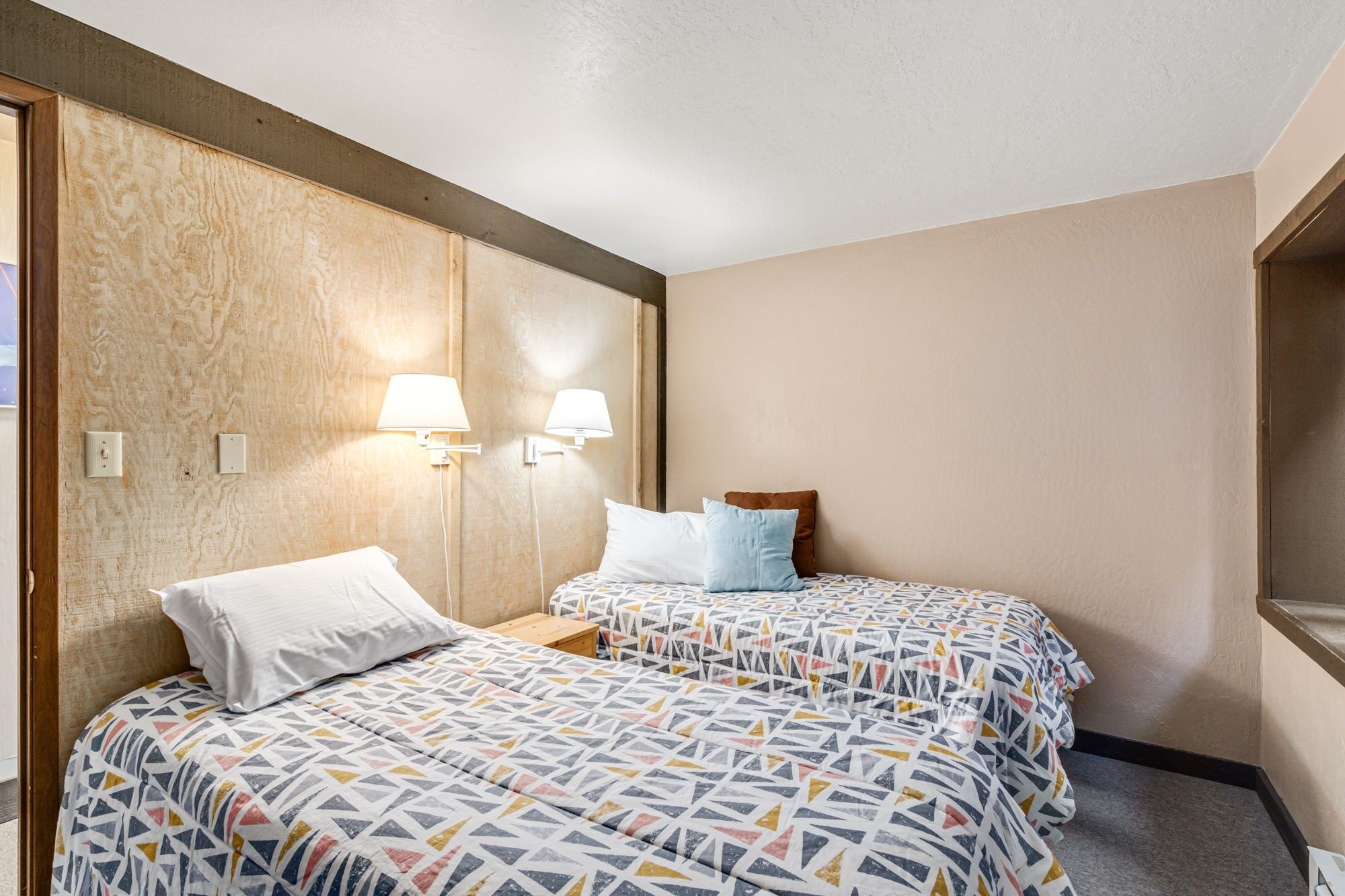 A hotel room with two beds, each with patterned bedding, a wooden headboard, and wall-mounted lamps.