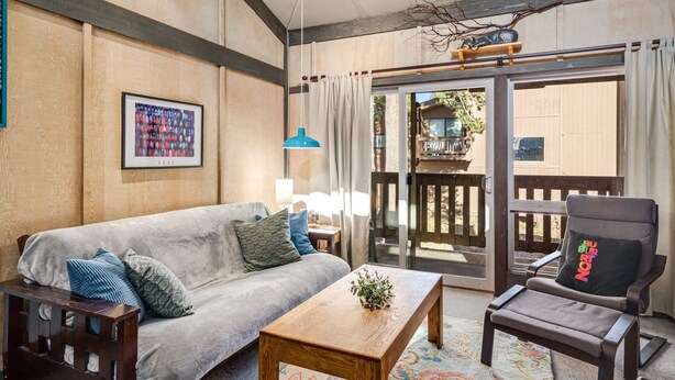 Una sala de estar acogedora con un sofá, una mesa de centro de madera y una silla. Hay un cuadro en la pared y una ventana con cortinas.