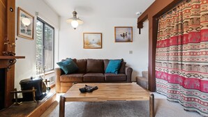 A cozy living room with a wooden coffee table, a brown sofa with blue cushions, and a large window. - Cozy, Well Furnished Condo! - Tamarack #12 by Bear Valley Vacation Rentals (Arnold)