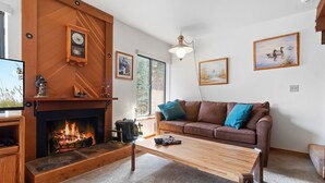 A cozy living room with a fireplace, a brown sofa with blue cushions, and framed artwork on the walls. - Cozy, Well Furnished Condo! - Tamarack #12 by Bear Valley Vacation Rentals (Arnold)