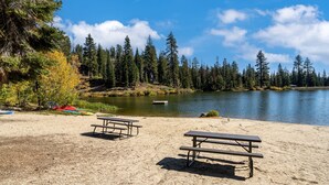 A sandy beach with two picnic tables and a lake surrounded by trees.