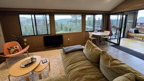 A modern living room with a large window, a flat-screen TV, a sofa, a small table, and a dining area. - Stunning! Enjoy The Best Views In All Of Bear Valley When You Stay At This Unique Property 3 Bedroom Apts by RedAwning (Arnold)