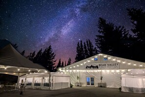 Un espace événementiel aménagé sous une tente, avec un ciel étoilé au-dessus, une tente de réception ornée d'une enseigne indiquant « Festival de musique de Bear Valley », et un paysage montagneux en arrière-plan.