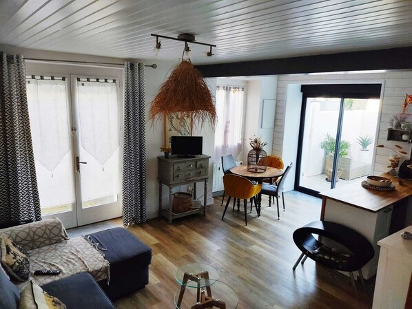 A modern living room with a sofa, ottoman, and a glass coffee table. There is a dining area with a table and chairs, and a kitchen area with a counter and cabinets.