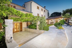 A courtyard with stone flooring, a white building with wooden shutters, and a well-maintained garden.
