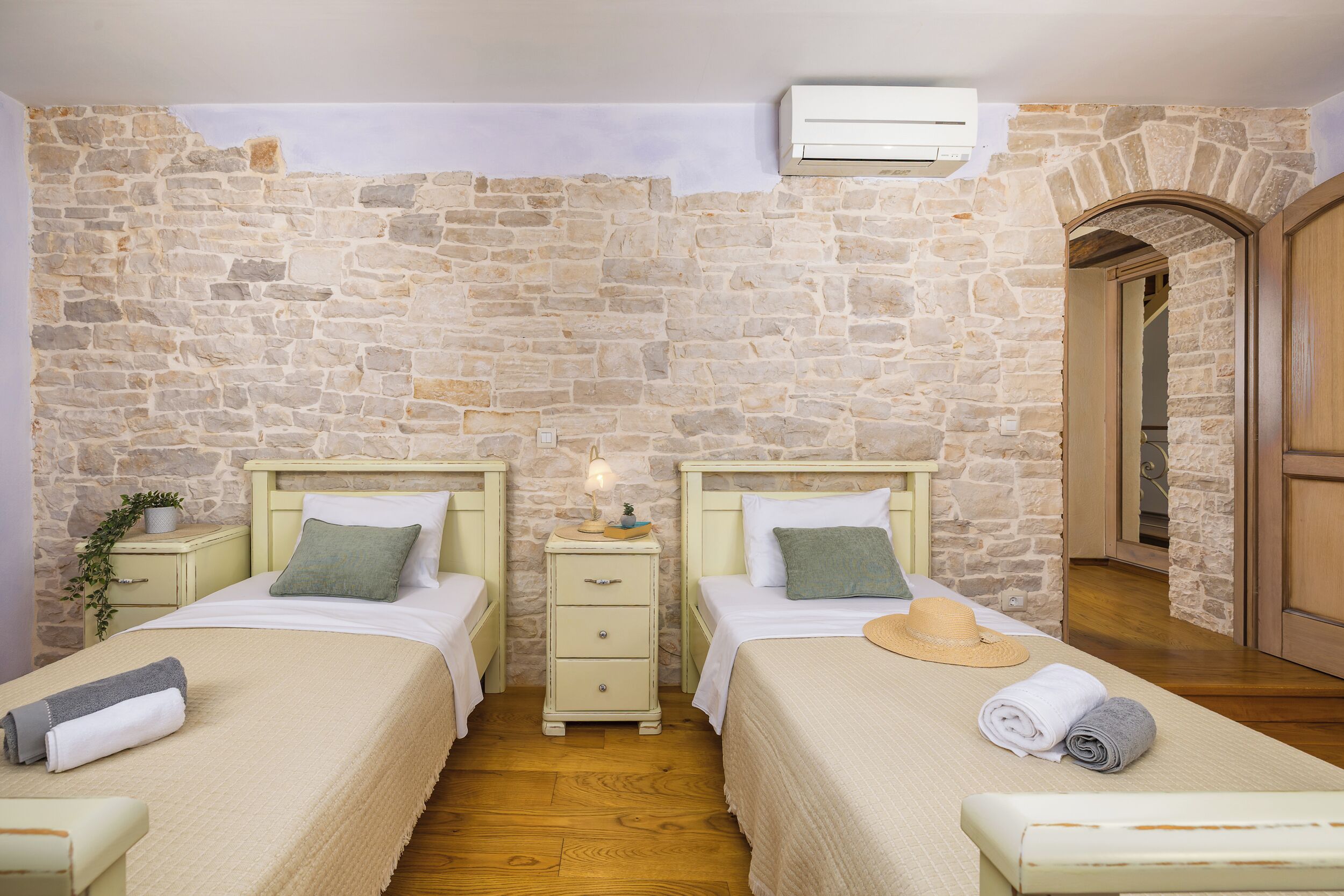 Une chambre avec deux lits, un mur en pierre, un plancher en bois, une table de chevet avec une lampe et un appareil de climatisation.