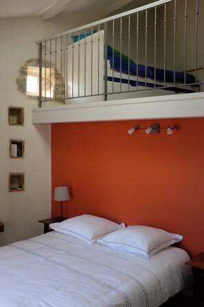 A bedroom with a bunk bed, a white bed with two pillows, a nightstand with a lamp, and a wall-mounted shelf with books. - Restored dovecote (Ardoix)