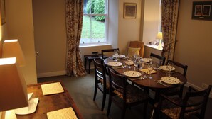 Comedor con una mesa redonda dispuesta para cuatro personas, rodeada de sillas. Se observa una ventana con cortinas, una lámpara a la izquierda y una pequeña mesa con una lámpara y adornos a la derecha.