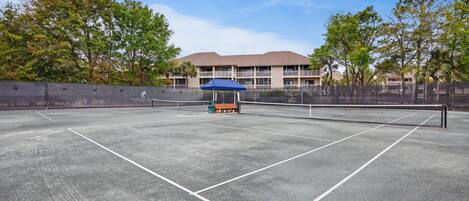 Una cancha de tenis con una red, una sombrilla azul y un edificio al fondo.