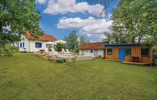 A rustic house with a red-tiled roof, a small wooden structure with a blue door, and a grassy yard with outdoor seating.