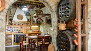 A rustic kitchen with stone walls, wooden beams, and a stone floor. There are wooden barrels, a hanging basket, and a table with chairs. - Once Upon a Time - A Castle of Magical Sunsets (Kalamata)