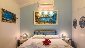 Quarto com cama, duas mesas de cabeceira, ar condicionado fixado na parede e um quadro de uma paisagem costeira.