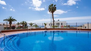 A clear blue swimming pool with a tiled edge, surrounded by a metal fence, palm trees, and a building with a satellite dish in the background. - Bungalow apartment with private pool & ocean views (Adeje)