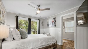 Une chambre à coucher avec un lit, un ventilateur de plafond, une fenêtre avec des rideaux, un tableau au mur et une vue sur l’extérieur.