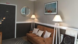 Quarto com sofá marrom, duas luminárias de chão, um quadro emoldurado e uma porta com a inscrição '101'.