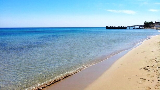 Une plage à l’eau claire, avec un rivage sablonneux et une jetée qui s’avance dans l’eau.