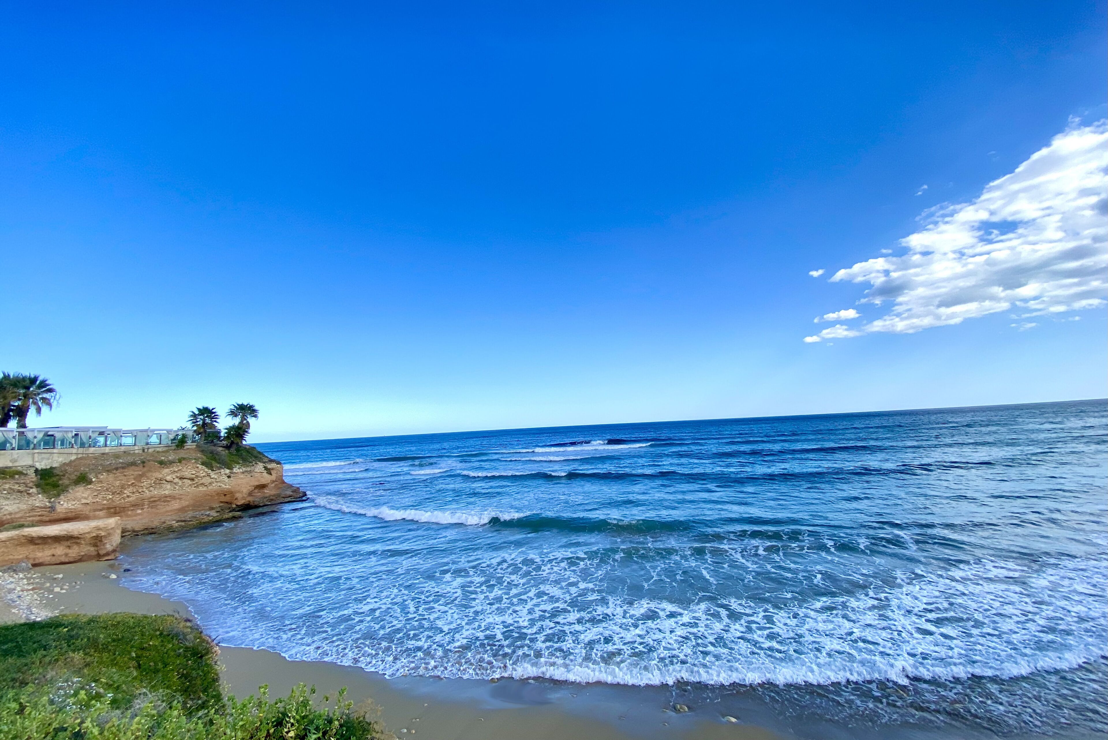 A coastal view with a clear sky, waves, and a structure with palm trees on a cliff.