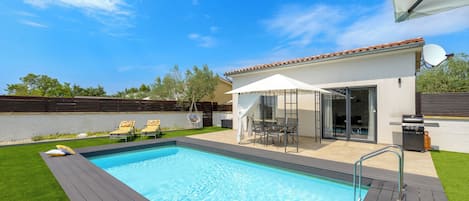 Une piscine avec une terrasse, un patio couvert aménagé avec du mobilier d’extérieur et une vue sur le ciel.