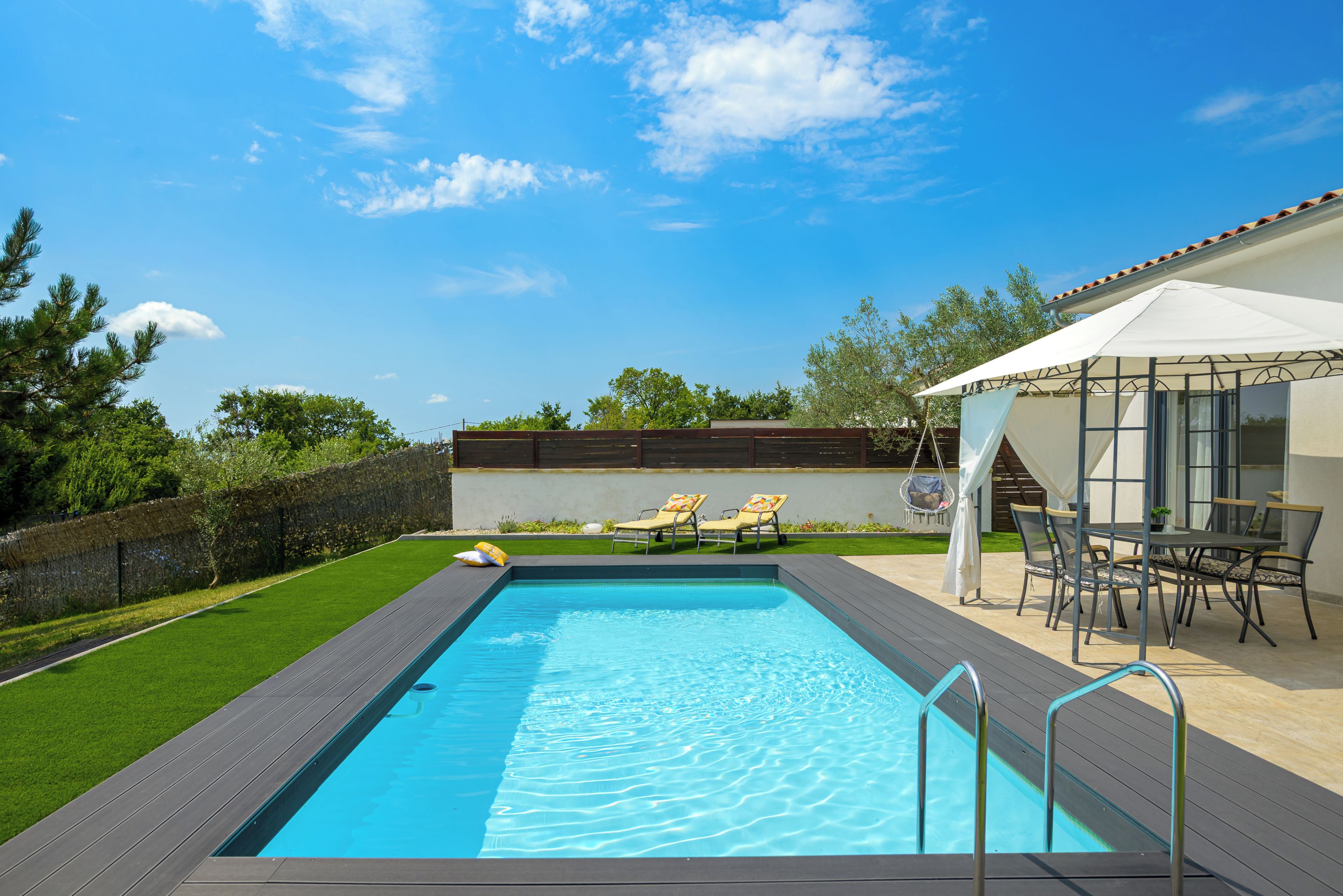 A rectangular swimming pool with a wooden deck, surrounded by a well-maintained lawn and a stone wall.