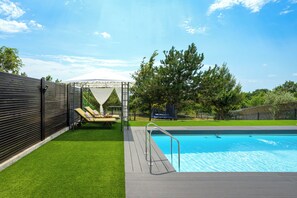 Une piscine avec une terrasse en bois, un espace salon couvert et un jardin clôturé.