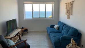 A living room with a large window overlooking the sea, a blue sofa, a TV on a wooden stand, and a macrame wall hanging. - Casa do Porto, relax with this stunning sea view (Porto Formoso)