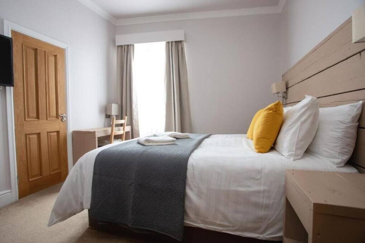 A bedroom with a bed, a wooden headboard, a nightstand, a lamp, a window with curtains, and a door.