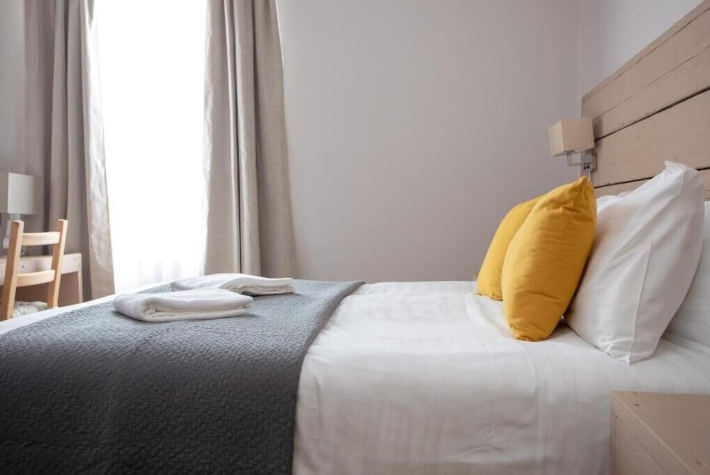 Una cama bien hecha con almohadas blancas y amarillas, una manta gris y una toalla doblada al lado.