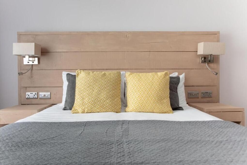 A neatly made bed with a grey blanket, two pillows with a yellow geometric pattern, and a wooden headboard.
