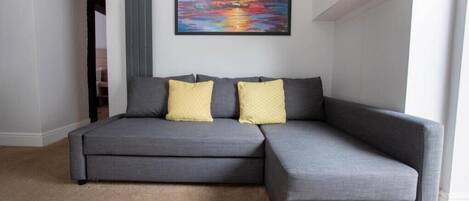 Un sofá seccional gris con dos cojines amarillos, un cuadro de un atardecer sobre el agua en la pared y una alfombra color beige.