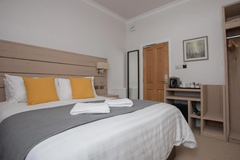 Una cama bien hecha con almohadas blancas y amarillas, un edredón gris y toallas blancas. Se aprecia un cabecero de madera, una mesita de noche con una lámpara y un armario con espejo.