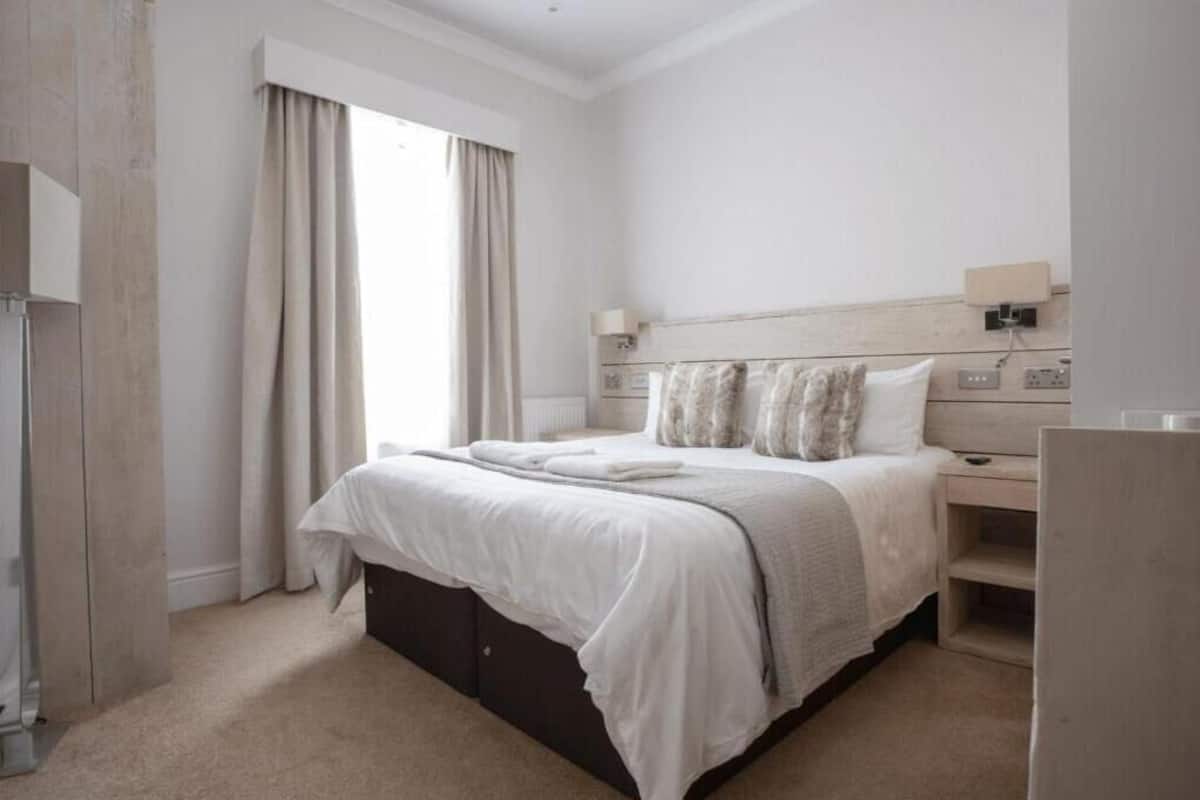 A modern hotel room with a large bed, a bedside table, a nightstand, a lamp, and a window with curtains.
