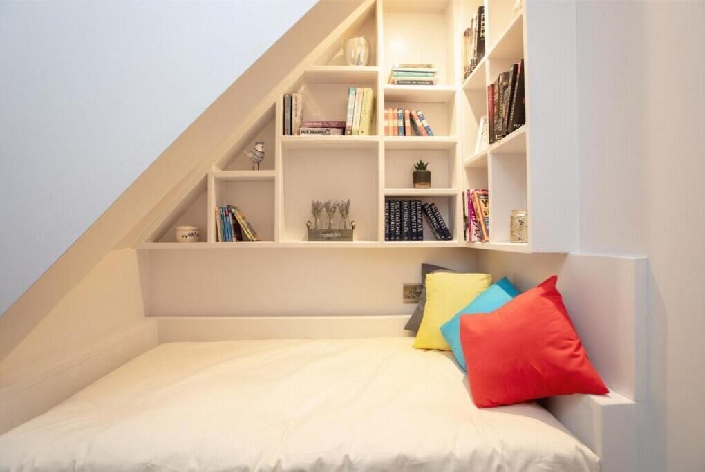 A bedroom with a sloped ceiling, a bed with white bedding, and a built-in bookshelf with books and decorative items.