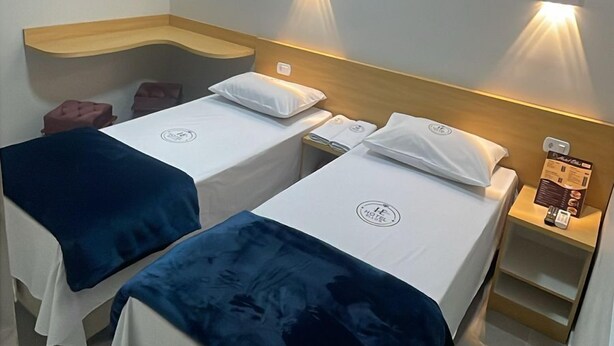 A hotel room with two single beds, each with a blue and white bedspread, and a wooden headboard. There are wall-mounted lights on either side of the bed, and a small wooden shelf with a menu on the right side. - Hotel Ellos (Itaquaquecetuba)