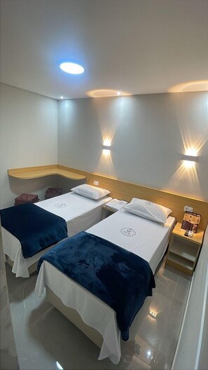 A hotel room with two single beds, each with a blue and white bedspread, and a wooden headboard. There are wall-mounted lights on either side of the bed, and a small wooden shelf with a menu on the right side. - Hotel Ellos (Itaquaquecetuba)