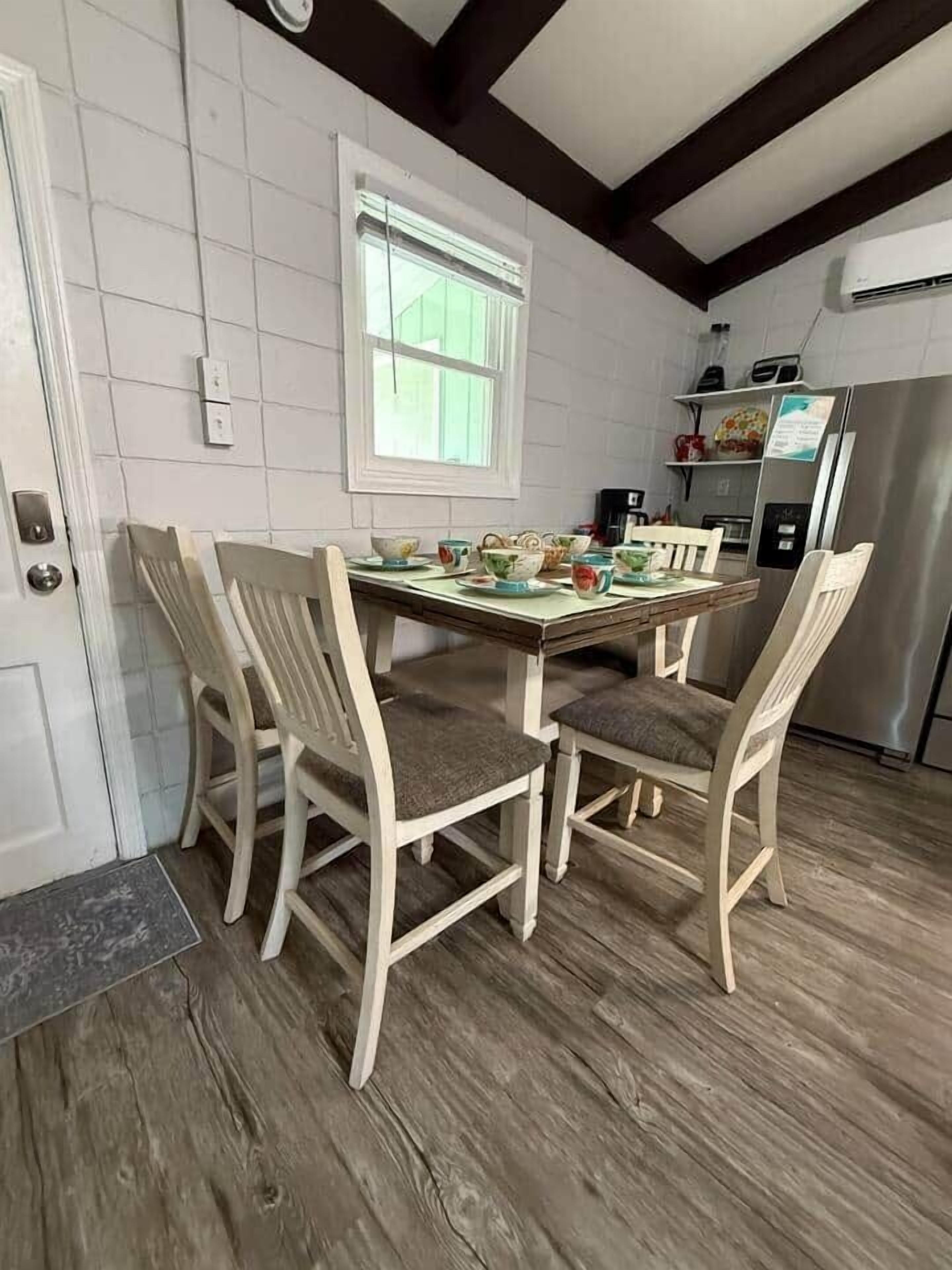 A dining area with a wooden table and chairs, a window, and a kitchen in the background.