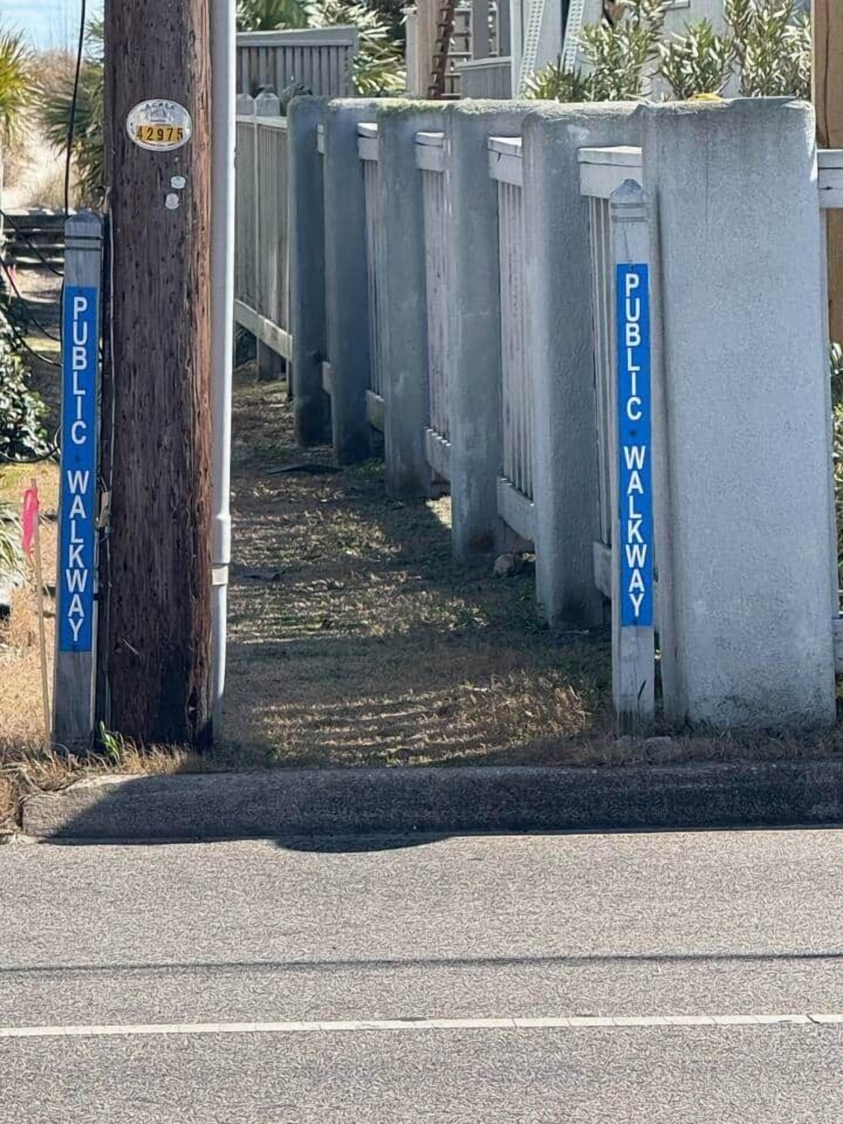 Un chemin public, accessible par une porte, bordé d’une clôture en bois et agrémenté d’un poteau électrique sur lequel est fixée une signalisation.