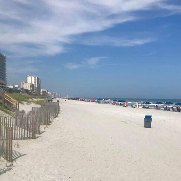 Une plage avec une clôture en bois, une poubelle et des bâtiments en arrière-plan.