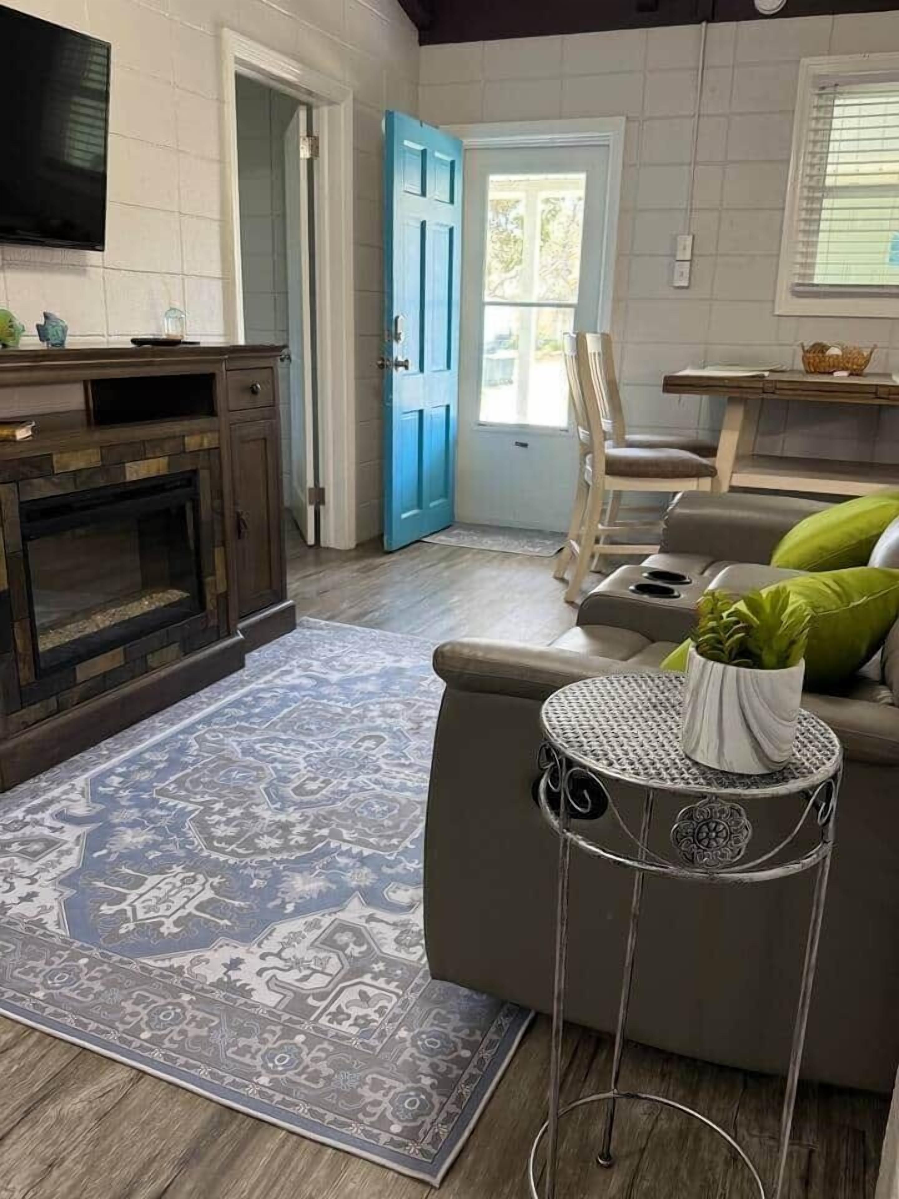 Una sala de estar con chimenea, una puerta azul, un sofá con cojines verdes y una pequeña mesa redonda con una planta.