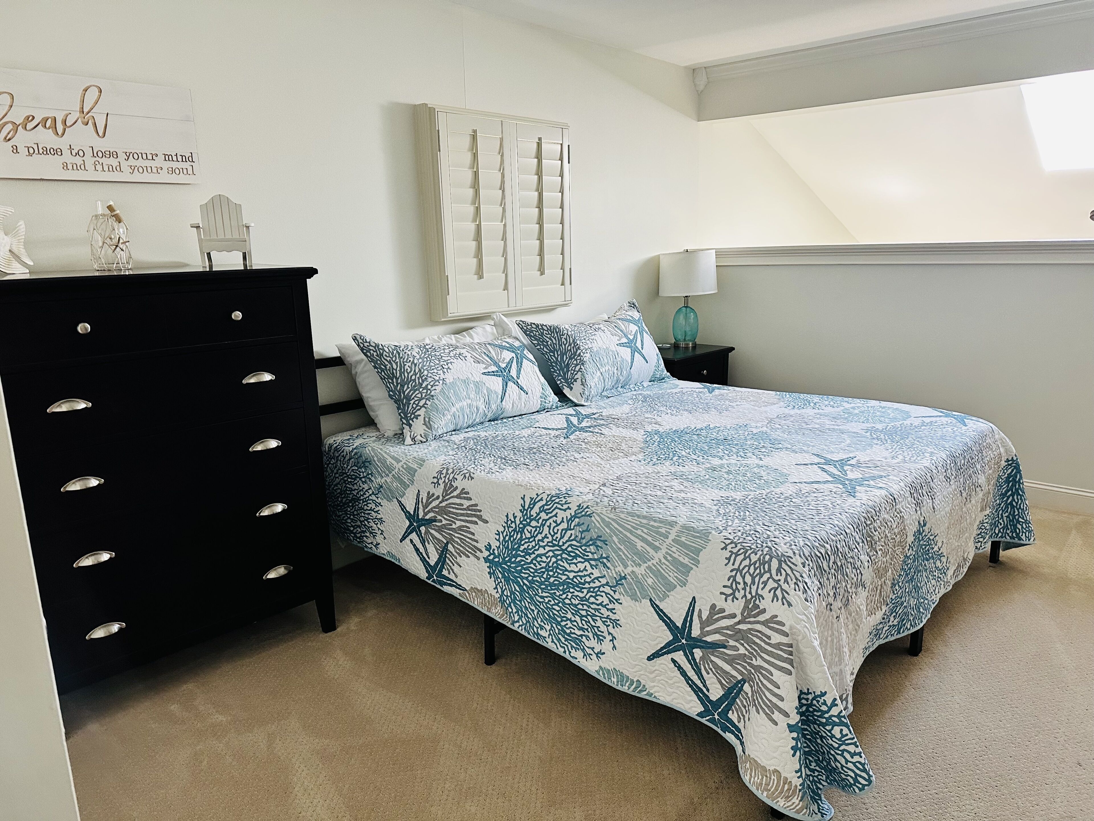Een slaapkamer met een bed, een dressoir, een nachtkastje, een lamp, een raam en een muur met een decoratief bord.
