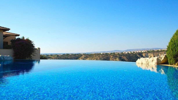 Una piscina infinita de color azul intenso con vistas a colinas distantes y un edificio a la izquierda.
