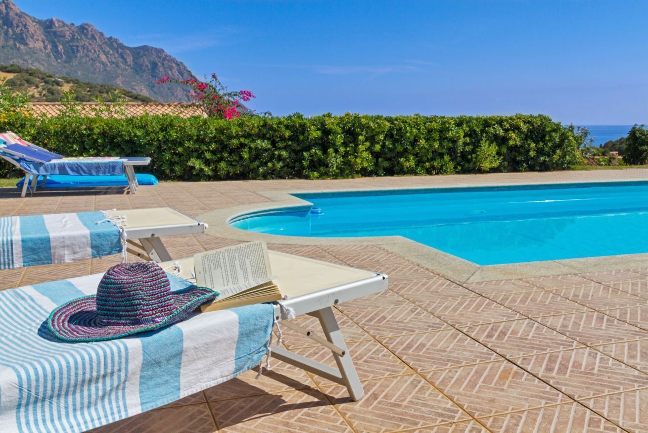 A poolside area with lounge chairs, a hat, and a book, surrounded by a tiled patio and a view of the sea and mountains.