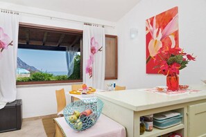 Ein Zimmer mit Esstisch, Stühlen, einer Obstschale, einer Vase mit Blumen, einem Gemälde und Blick aufs Meer.
