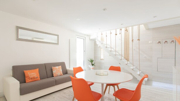 A modern living room with a white sofa, orange cushions, and a round table with orange chairs. A staircase with a white railing and decorative rope lights is visible in the background.